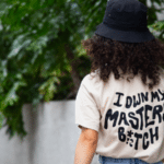 Person with curly hair wearing a black bucket hat and a beige shirt with the phrase "I own my master b*tch" printed on the back, standing in front of green foliage; perfect merch for making a bold statement.