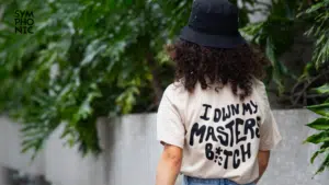 Person with curly hair wearing a black bucket hat and a beige shirt with the phrase "I own my master b*tch" printed on the back, standing in front of green foliage; perfect merch for making a bold statement.