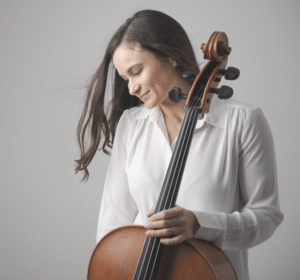 A person with long brown hair is holding a cello, wearing a white shirt, and looking down with an unfiltered slight smile.