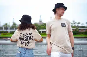 Two people in casual attire stand by a railing. The person on the left wears a hat and a shirt that reads "I own my masters b*tch," and the person on the right wears a hat and a "Symphonic" shirt.