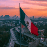 A large Mexican flag is prominently displayed with an elevated highway and busy traffic below, set against a cityscape and an orange sunset sky in the background.