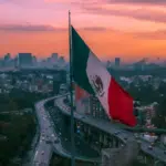 A large Mexican flag is prominently displayed with an elevated highway and busy traffic below, set against a cityscape and an orange sunset sky in the background.