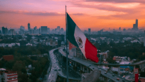 A large Mexican flag is prominently displayed with an elevated highway and busy traffic below, set against a cityscape and an orange sunset sky in the background.