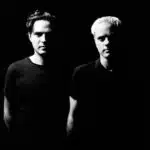 A black-and-white photo of two men standing side by side against a black background, partially illuminated. They are both wearing dark shirts. The man on the left has dark hair, and the man on the right has light hair, appearing like artists ready to debut their new music.