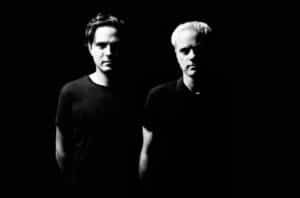 A black-and-white photo of two men standing side by side against a black background, partially illuminated. They are both wearing dark shirts. The man on the left has dark hair, and the man on the right has light hair, appearing like artists ready to debut their new music.