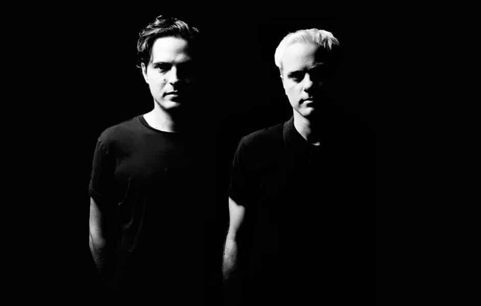 A black-and-white photo of two men standing side by side against a black background, partially illuminated. They are both wearing dark shirts. The man on the left has dark hair, and the man on the right has light hair, appearing like artists ready to debut their new music.