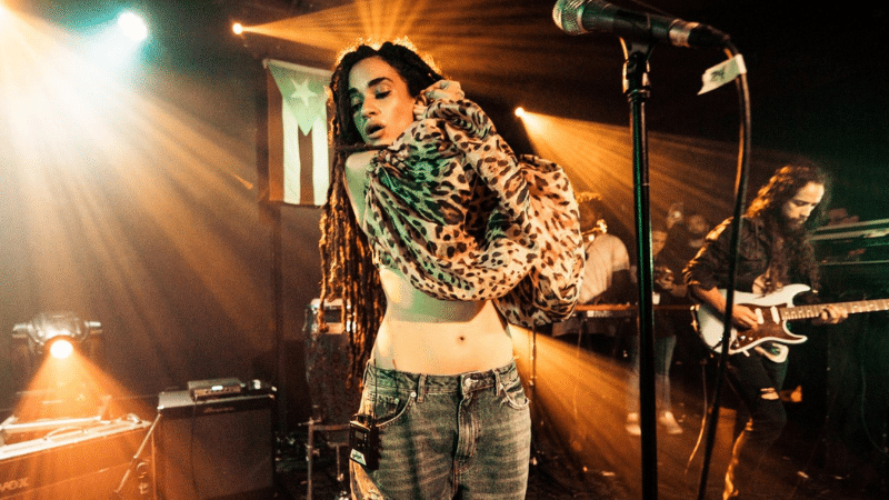 A singer on stage wearing jeans and a leopard-print top dances next to a guitarist under warm stage lights, with a microphone stand in the foreground.