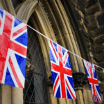 Bunting with multiple Union Jack flags strung along the exterior of a stone building with arched windows.