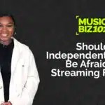 A person in a white jacket stands against a dark background with text: "MUSIC BIZ 101: Should Independent Artists Be Afraid of Streaming Fraud?