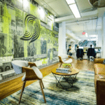 A modern office lounge area features a wall mural, two chairs, a coffee table with books, a brown sofa, and a blue rug. People work at desks in the background. A "Symphonic" sign is visible.