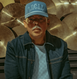 Man wearing a blue "Local" cap and denim jacket sits against a backdrop of metallic discs.