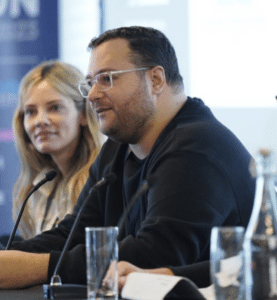 Two individuals are seated at a panel discussion table with microphones; one person is speaking while the other listens attentively.