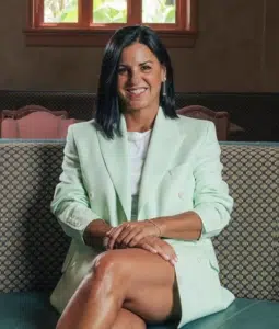 A woman with shoulder-length dark hair wearing a light green blazer sits on a patterned couch with her legs crossed, smiling at the camera.