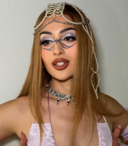 A person with colorful makeup and long hair adorned with a pearl headpiece, wearing glasses and a choker necklace, poses against a plain background.