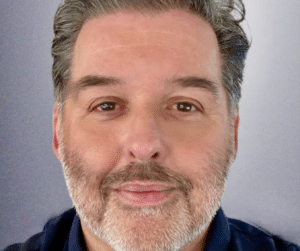 A close-up of a middle-aged man with short grey hair and a beard, looking directly at the camera against a plain background.