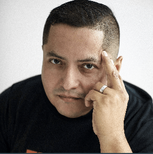 A man with short dark hair and a black shirt, wearing a ring on his left ring finger, rests his fingers on the side of his forehead. He has a neutral facial expression and looks directly at the camera.