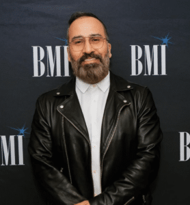 A person with a beard and glasses wearing a leather jacket standing in front of a backdrop with "BMI" printed on it.