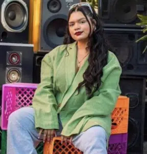 A person with long, dark hair, wearing a green oversized jacket and grey sweatpants, sits on colorful plastic crates in front of large audio speakers, clearly immersed in the music.