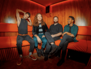 Four men sit on a red cushioned bench in a room with wooden paneling. Three are seated casually while one stretches his arms above his head. They all appear relaxed and are smiling or laughing.