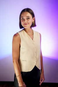 A person with short brown hair and wearing a beige vest smiles at the camera against a purple gradient background.