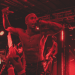 A shirtless man performs energetically on stage with a microphone while others dance in the background under red lighting.