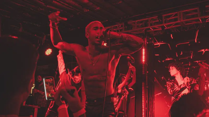 A shirtless man performs energetically on stage with a microphone while others dance in the background under red lighting.