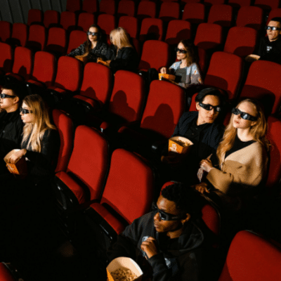 People wearing 3D glasses sit in a movie theater with red seats, some holding popcorn. In perfect sync, they all lean forward during an intense scene.