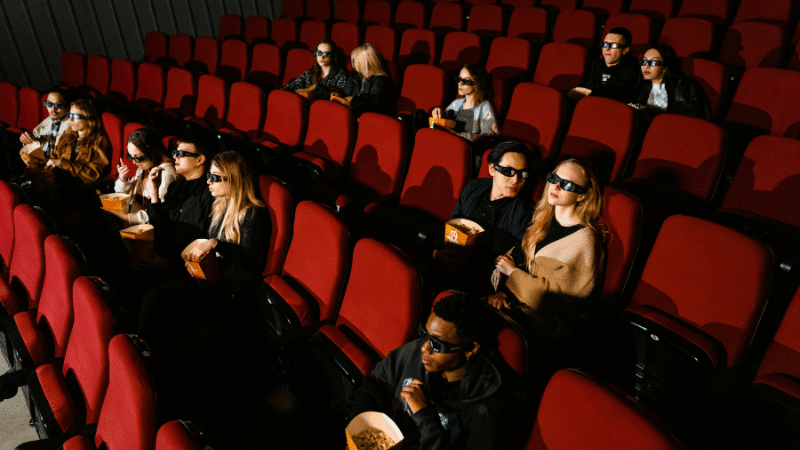 People wearing 3D glasses sit in a movie theater with red seats, some holding popcorn. In perfect sync, they all lean forward during an intense scene.