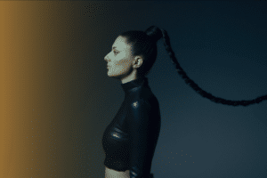 A person in profile with a long braided ponytail, dressed in a black high-neck top, stands against a gradient background transitioning from yellow to dark gray, exuding the vibe of new music.