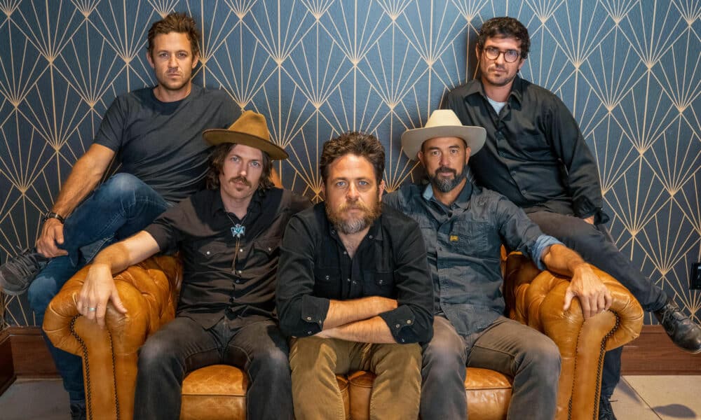 Five men are seated on or standing behind a leather couch in front of a patterned background. Three are wearing hats and all are dressed in casual attire, perhaps gearing up to discuss their new music.