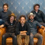 Five men are seated on or standing behind a leather couch in front of a patterned background. Three are wearing hats and all are dressed in casual attire, perhaps gearing up to discuss their new music.