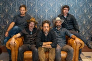 Five men are seated on or standing behind a leather couch in front of a patterned background. Three are wearing hats and all are dressed in casual attire, perhaps gearing up to discuss their new music.