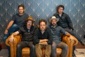 Five men are seated on or standing behind a leather couch in front of a patterned background. Three are wearing hats and all are dressed in casual attire, perhaps gearing up to discuss their new music.