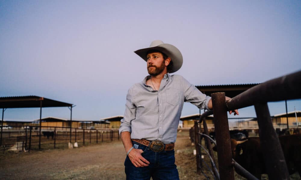 A man wearing a cowboy hat and casual clothing stands by a fence in a ranch setting, looking into the distance. Ranch buildings and cattle are visible in the background, while the tunes of new music play softly, adding a modern touch to the rustic scene.