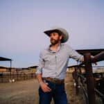 A man wearing a cowboy hat and casual clothing stands by a fence in a ranch setting, looking into the distance. Ranch buildings and cattle are visible in the background, while the tunes of new music play softly, adding a modern touch to the rustic scene.