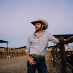 A man wearing a cowboy hat and casual clothing stands by a fence in a ranch setting, looking into the distance. Ranch buildings and cattle are visible in the background, while the tunes of new music play softly, adding a modern touch to the rustic scene.