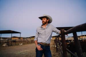 A man wearing a cowboy hat and casual clothing stands by a fence in a ranch setting, looking into the distance. Ranch buildings and cattle are visible in the background, while the tunes of new music play softly, adding a modern touch to the rustic scene.