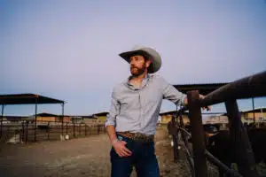 A man wearing a cowboy hat and casual clothing stands by a fence in a ranch setting, looking into the distance. Ranch buildings and cattle are visible in the background, while the tunes of new music play softly, adding a modern touch to the rustic scene.