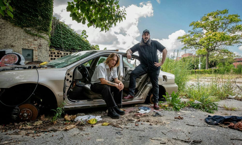 Two individuals pose by a stripped car in an overgrown, graffiti-marked area. One sits on the car seat, while the other stands leaning against the open door, discussing new music. The sky is partially cloudy.