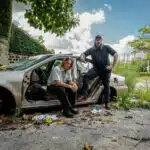 Two individuals pose by a stripped car in an overgrown, graffiti-marked area. One sits on the car seat, while the other stands leaning against the open door, discussing new music. The sky is partially cloudy.