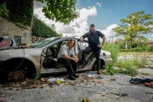 Two individuals pose by a stripped car in an overgrown, graffiti-marked area. One sits on the car seat, while the other stands leaning against the open door, discussing new music. The sky is partially cloudy.