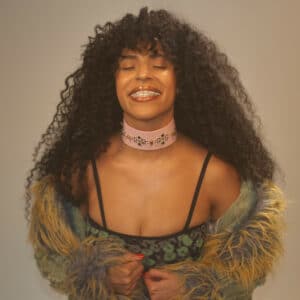 A woman with long curly hair and closed eyes smiles while holding her coat. She wears a pink choker and a patterned top with straps.