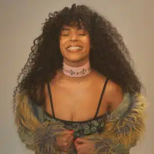 A woman with long curly hair and closed eyes smiles while holding her coat. She wears a pink choker and a patterned top with straps.