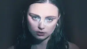 Close-up of a person with wet hair, wearing pearls on their face and large hoop earrings, gazing directly at the camera—a case study in artistic expression. The background is dark and blurred.