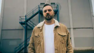 A man in a beige jacket stands in front of a building with an exterior staircase.