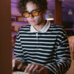 In a room aglow with dim purple lighting, a person in glasses and a striped shirt types intently on a keyboard, as the glow from the screen seems to rise and illuminate their focused expression.