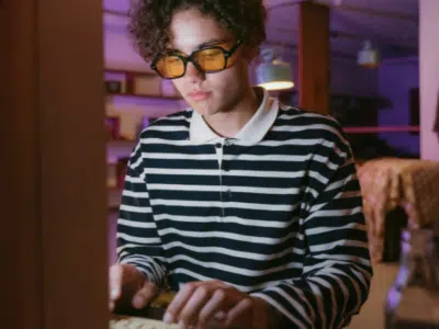 In a room aglow with dim purple lighting, a person in glasses and a striped shirt types intently on a keyboard, as the glow from the screen seems to rise and illuminate their focused expression.