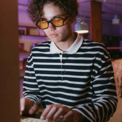 In a room aglow with dim purple lighting, a person in glasses and a striped shirt types intently on a keyboard, as the glow from the screen seems to rise and illuminate their focused expression.