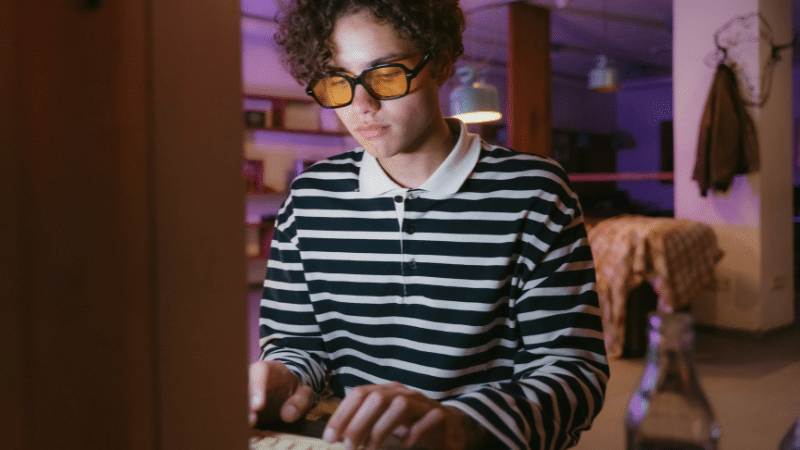 In a room aglow with dim purple lighting, a person in glasses and a striped shirt types intently on a keyboard, as the glow from the screen seems to rise and illuminate their focused expression.