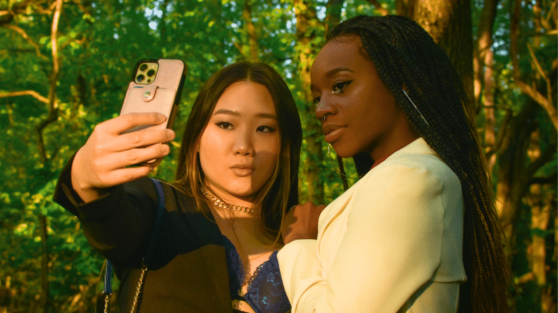 In a sunlit forest, two individuals pose for an elegant selfie, capturing their formal attire's sleek lines—perfect for sharing and boosting their Instagram analytics.